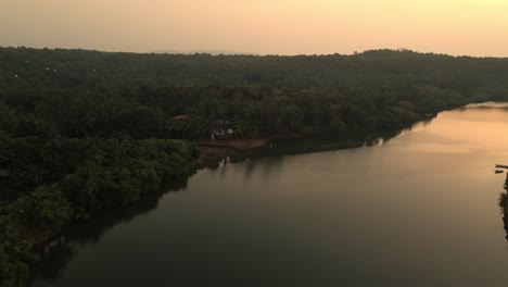 a serene drone shot of a calm river at sunset in goa, india, lush green forests on the both side under a glowing orange-golden sky, radiating peaceful beauty