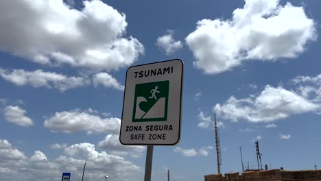 Primer-Plano-De-Un-Cartel-De-Tsunami-En-Una-Zona-Segura-Contra-El-Cielo-Azul-Y-Las-Nubes-En-El-Océano-En-Portugal