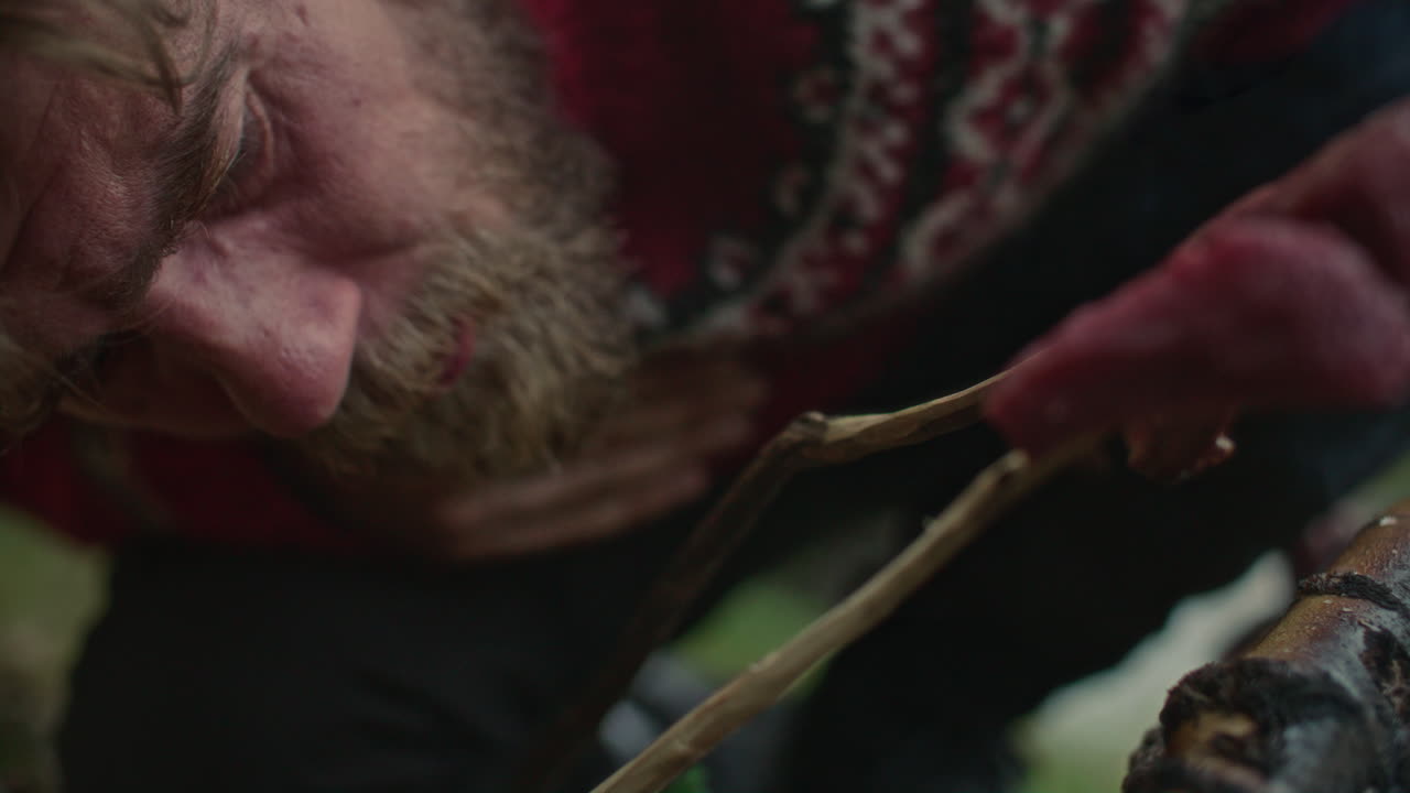 Man Blowing into Campfire while Cooking Meat in Wilderness