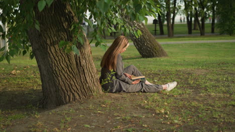 lady in grey clothing outdoors reading book with her hand on page, leaning against tree trunk, background features greenery with softly illuminated natural light