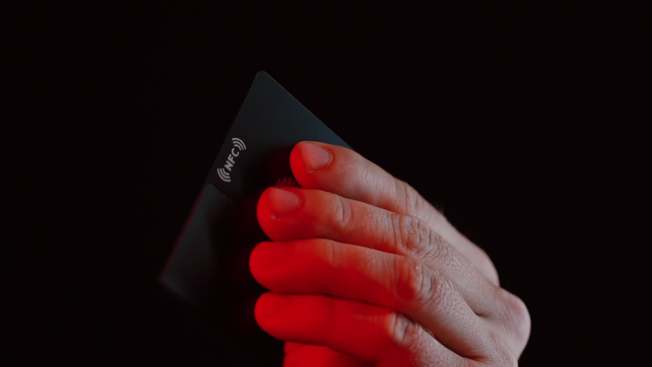 A close-up of a hand holding a black NFC card illuminated by red light in a dark background, symbolizing contactless technology, digital payments, and modern security concepts