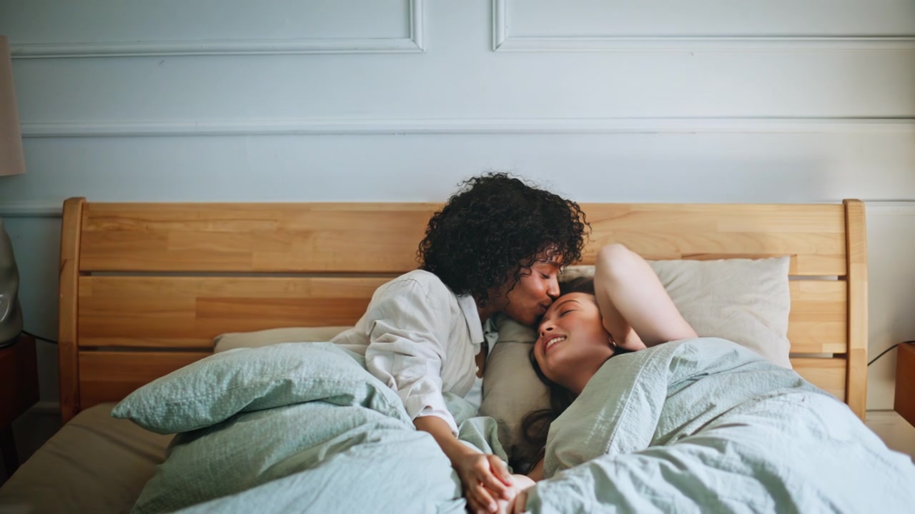 Lesbian couple cuddling bed morning. Two women holding hands, forehead kiss.