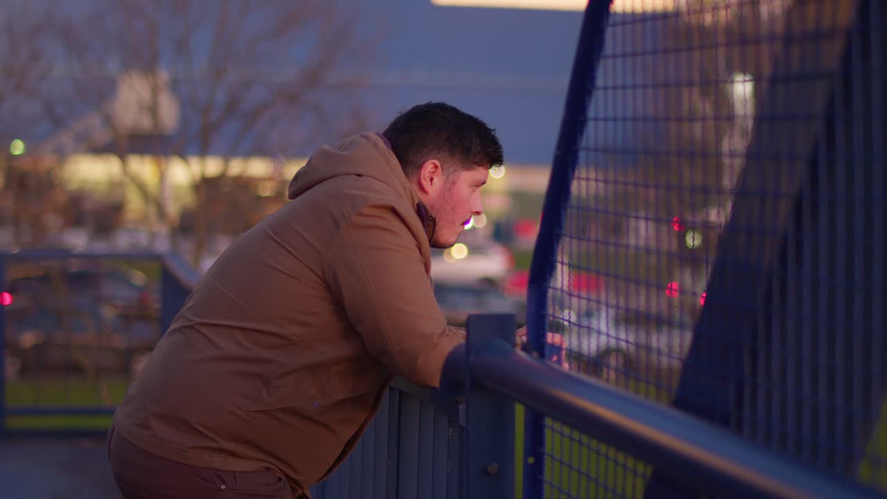 A medium shot of a man standing on a blue iron bridge, gazing into the distance with cars passing by in the blurred background. Slow-motion footage capturing a reflective, cinematic moment.