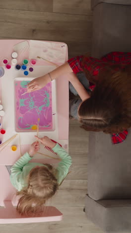 Woman teacher and little girl student make pattern with paints on oily water surface at Ebru art lesson in living room. Creative hobby upper view