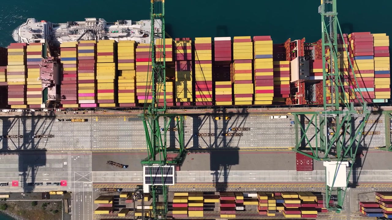 Cargo containers are stacked at a busy port. Cranes are positioned above the stacks. Delays in shipping are evident as the distribution of goods slows. Freight transportation challenges persist.