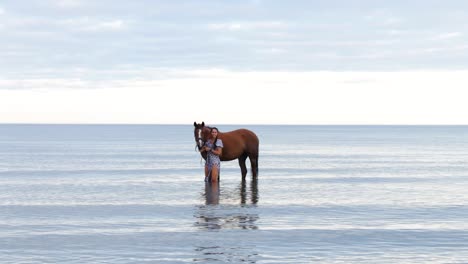 a beautiful girl with long hair in a blue dress posing with her horse in the water during the evening in donabate, ireland
