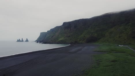 Rising-aerial-drone-shot-of-beautiful-blue-waves-crashing-on-a-black-sand-beach-in-Iceland-with-a-group-of-Icelandic-horses-and-huge-rock-formations-in-the-distance