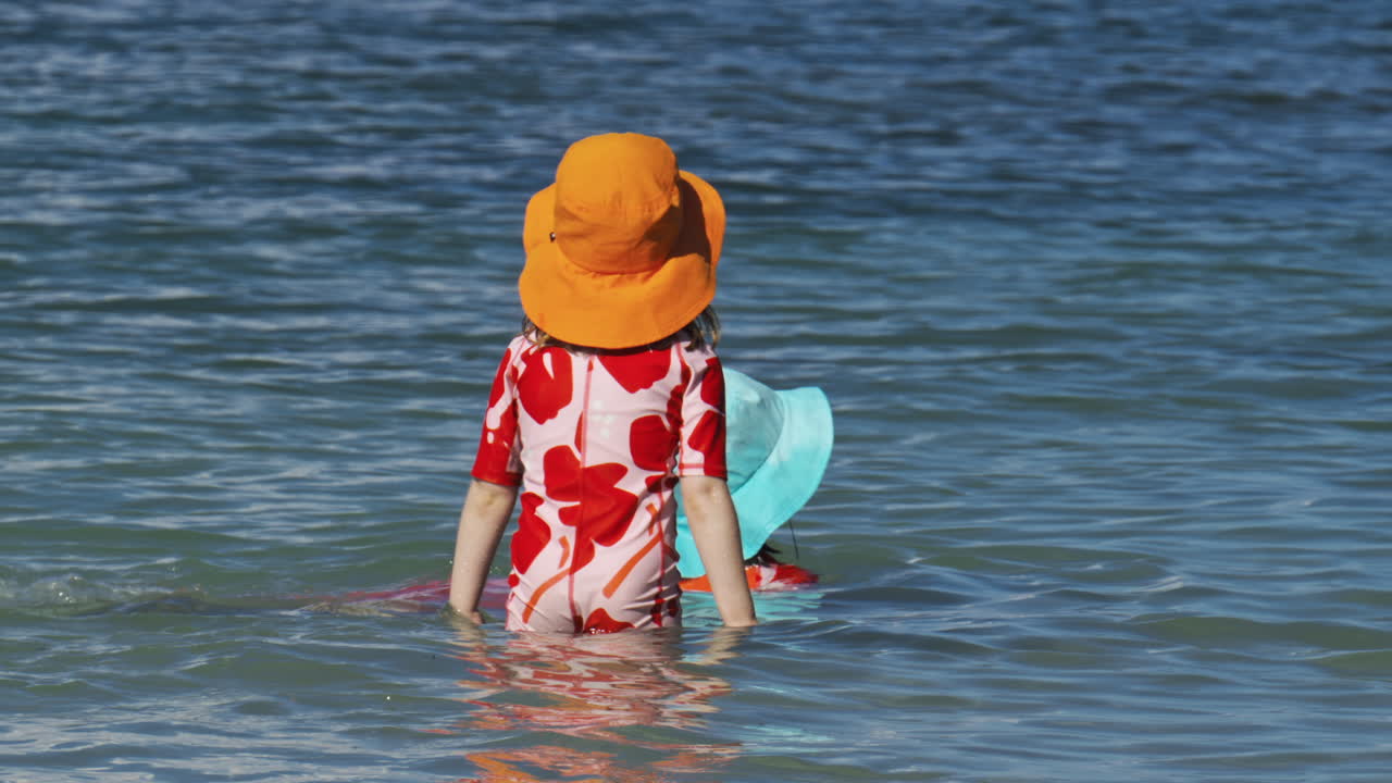 Little girl with an orange hat and a pink and red swimsuit walking through the sea