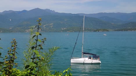 small sailboat lonley in a lake with mountains and forest in the background and green leaves in front