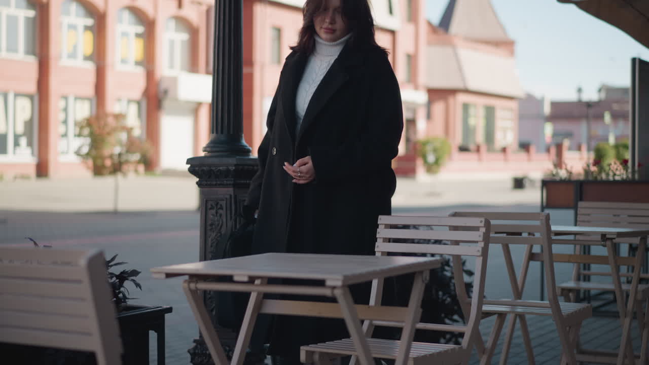 Businesswoman in black coat and white turtleneck sits down on outdoor restaurant chair, placing her bag on table with visible relief, urban background includes decorative poles, chairs and tables