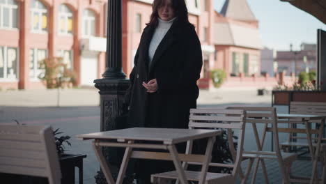 businesswoman in black coat and white turtleneck sits down on outdoor restaurant chair, placing her bag on table with visible relief, urban background includes decorative poles, chairs and tables