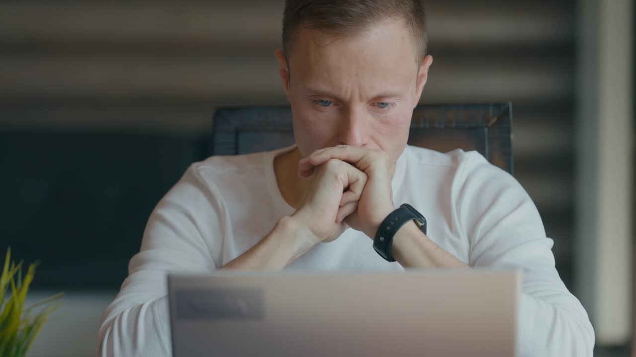 el hombre está mirando atentamente a la pantalla del portátil mientras está sentado en su escritorio en la habitación. sostiene las manos a la cara. mira pensativo hacia el lado