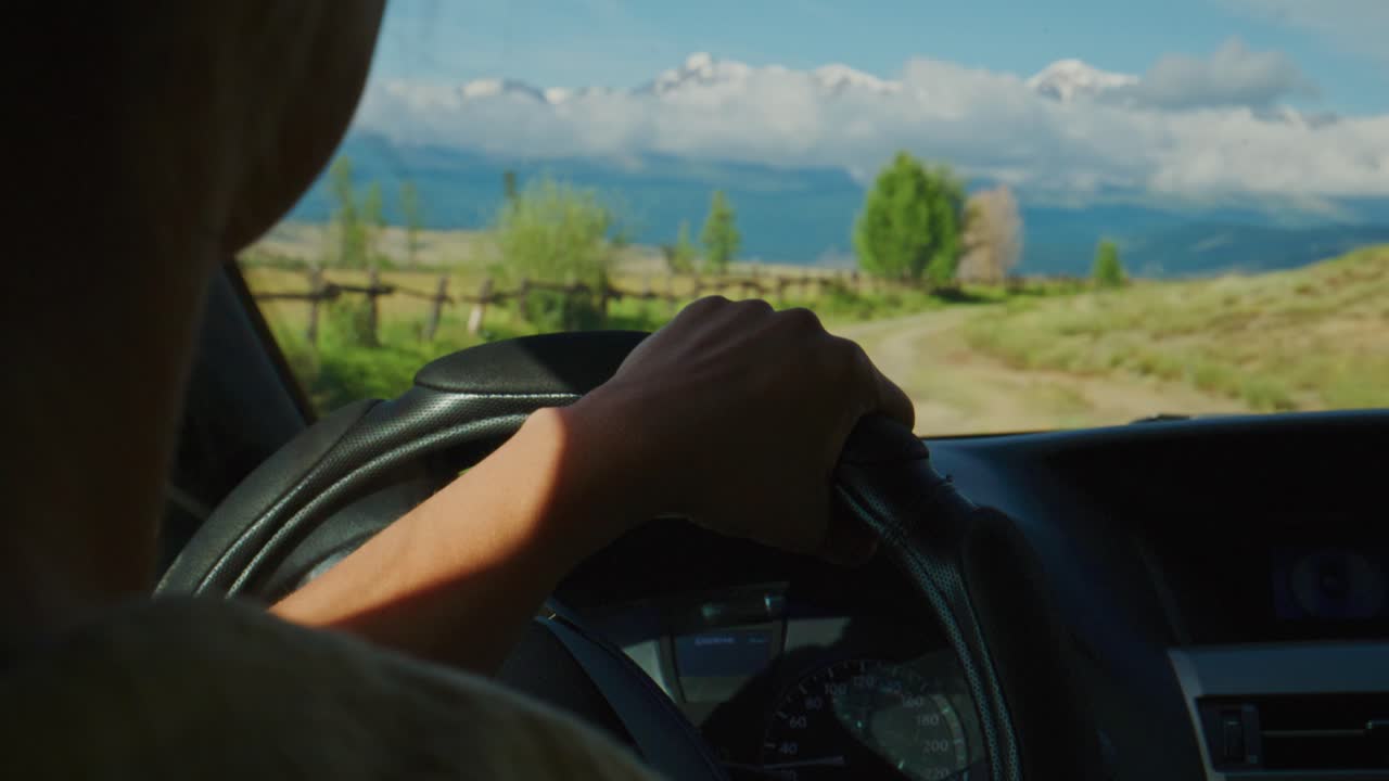 Mani del conducente che stringono il volante durante un viaggio su strada. La vista attraverso il parabrezza mostra campi soleggiati, una recinzione e montagne lontane. La persona è concentrata sulla strada da percorrere, calma e determinata. Ideale per viaggi e avventura.