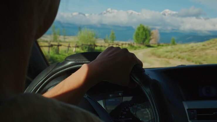 Mani del conducente che stringono il volante durante un viaggio su strada. La vista attraverso il parabrezza mostra campi soleggiati, una recinzione e montagne lontane. La persona è concentrata sulla strada da percorrere, calma e determinata. Ideale per viaggi e avventura.