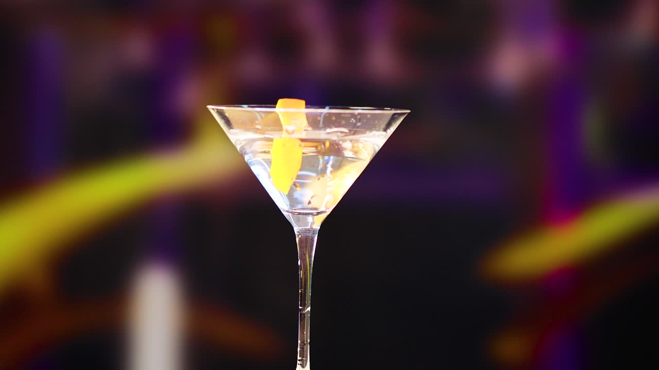 A martini glass with a lemon twist against a colorful, blurred background. Bright lighting highlights the glass and garnish