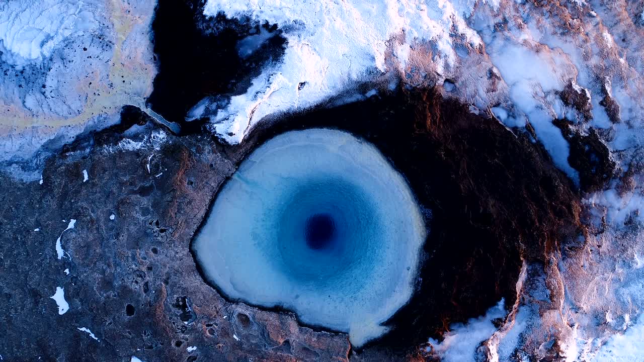 아이슬란드의 게이시르에 있는 게이저의 중심에서 점점 더 멀어지는 드론의 공중 사진. 이 게이저 밖으로 어나오고 있으며, 사람들은 은 해가 지는 곳에서 게이저를 둘러싸고 고 있습니다.