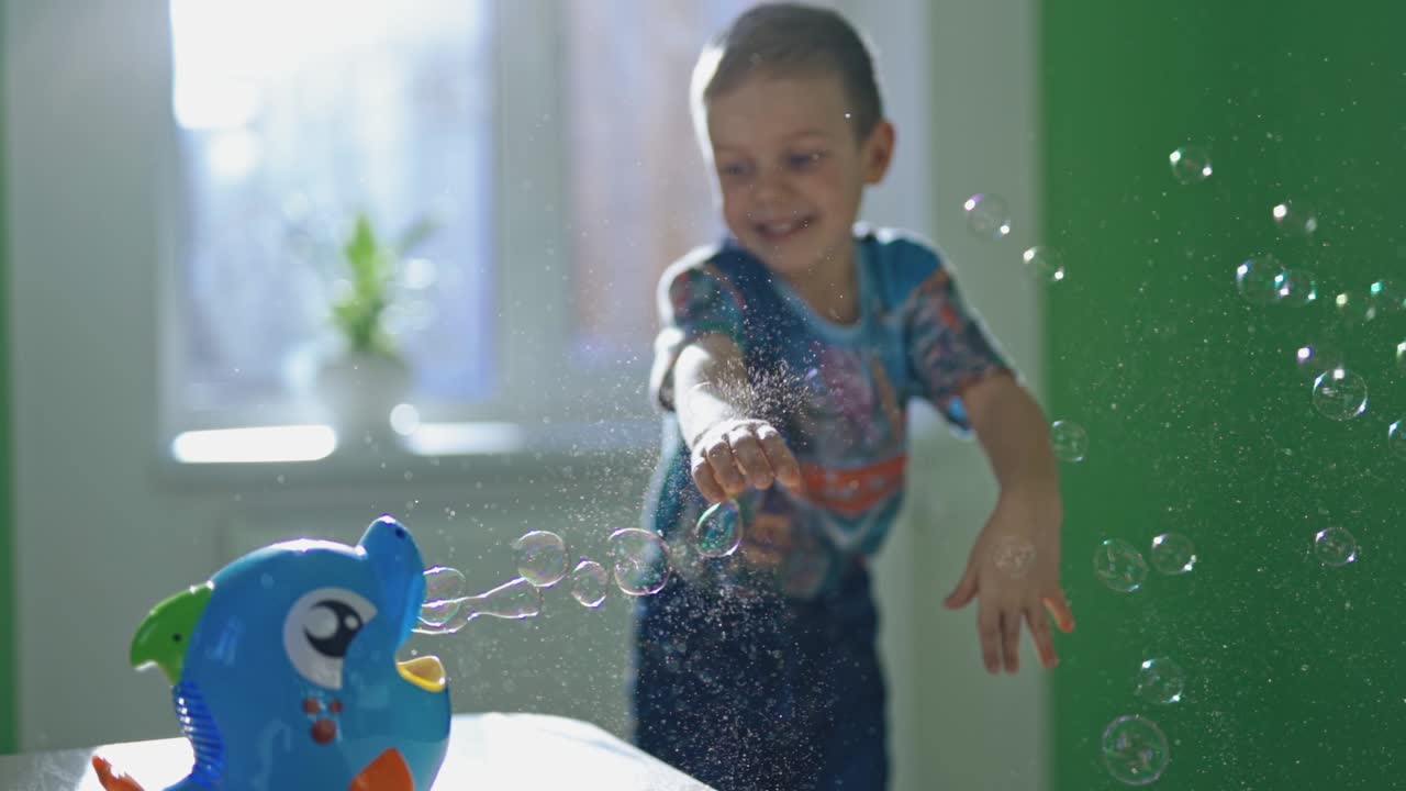 Little boy is popping soap bubbles indoors. Curious child catches circle bubbles at home. Bubble blower in the form of a fish and a small kid plays happily nearby.