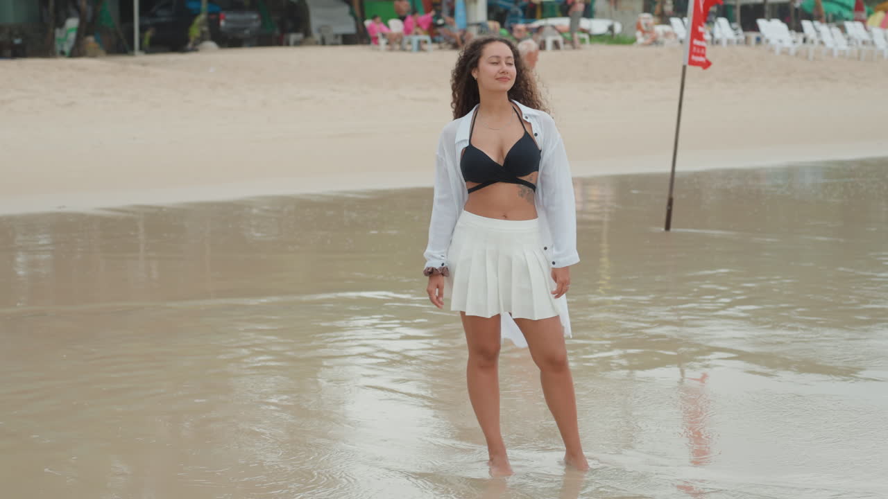 Modella in piedi sulla sabbia bagnata della riva di una spiaggia isolana, con top bikini nero, gonna bianca e camicia trasparente. L'acqua calma e poco profonda riflette il cielo nuvoloso. Posa rilassata, bagnanti in lontananza, onde delicate.