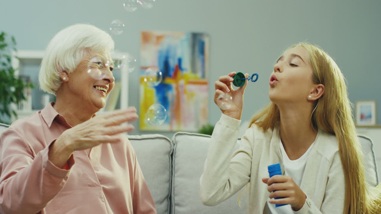 화려한 백인 할머니와 손녀가 거실의 소파에 함께 앉아 있고 소녀가 비누 거품을 불고 있는 초상화입니다. 실내.