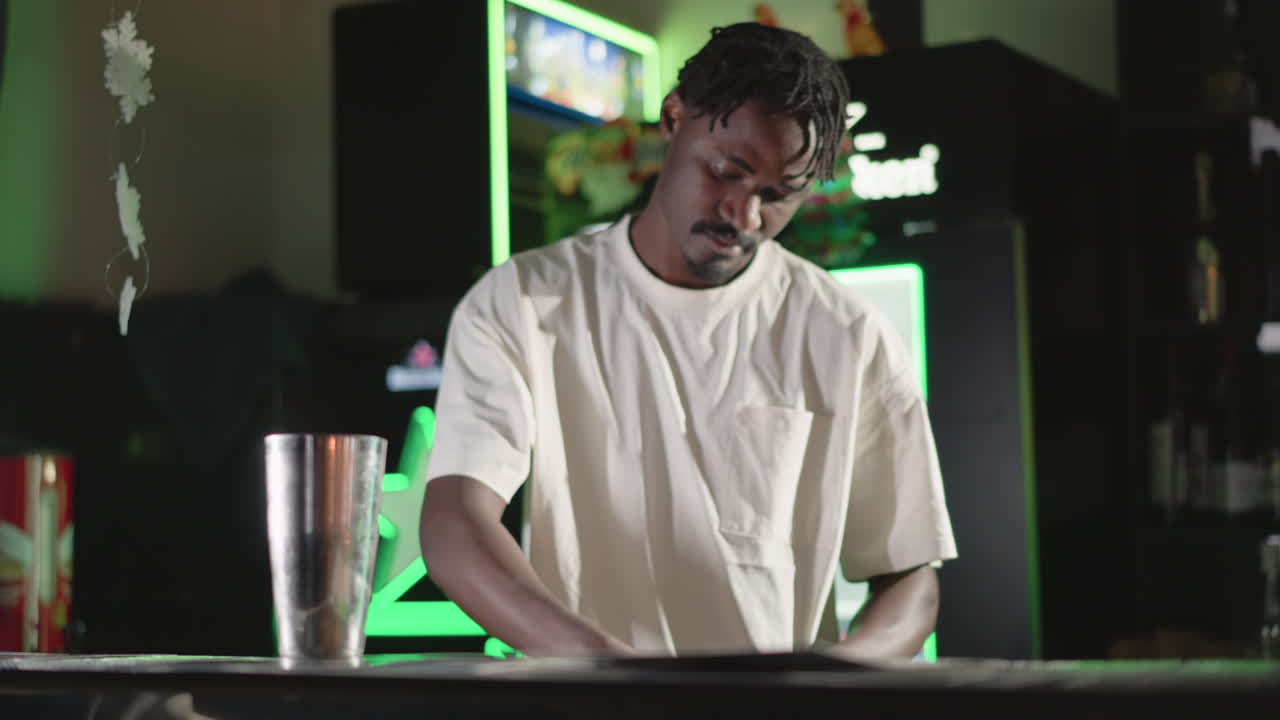 Guy preparing drinks in bar, holding glass in one hand and shaking cocktail shaker in other, focused on work with neon-lit bar background