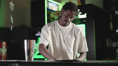 guy preparing drinks in bar, holding glass in one hand and shaking cocktail shaker in other, focused on work with neon-lit bar background