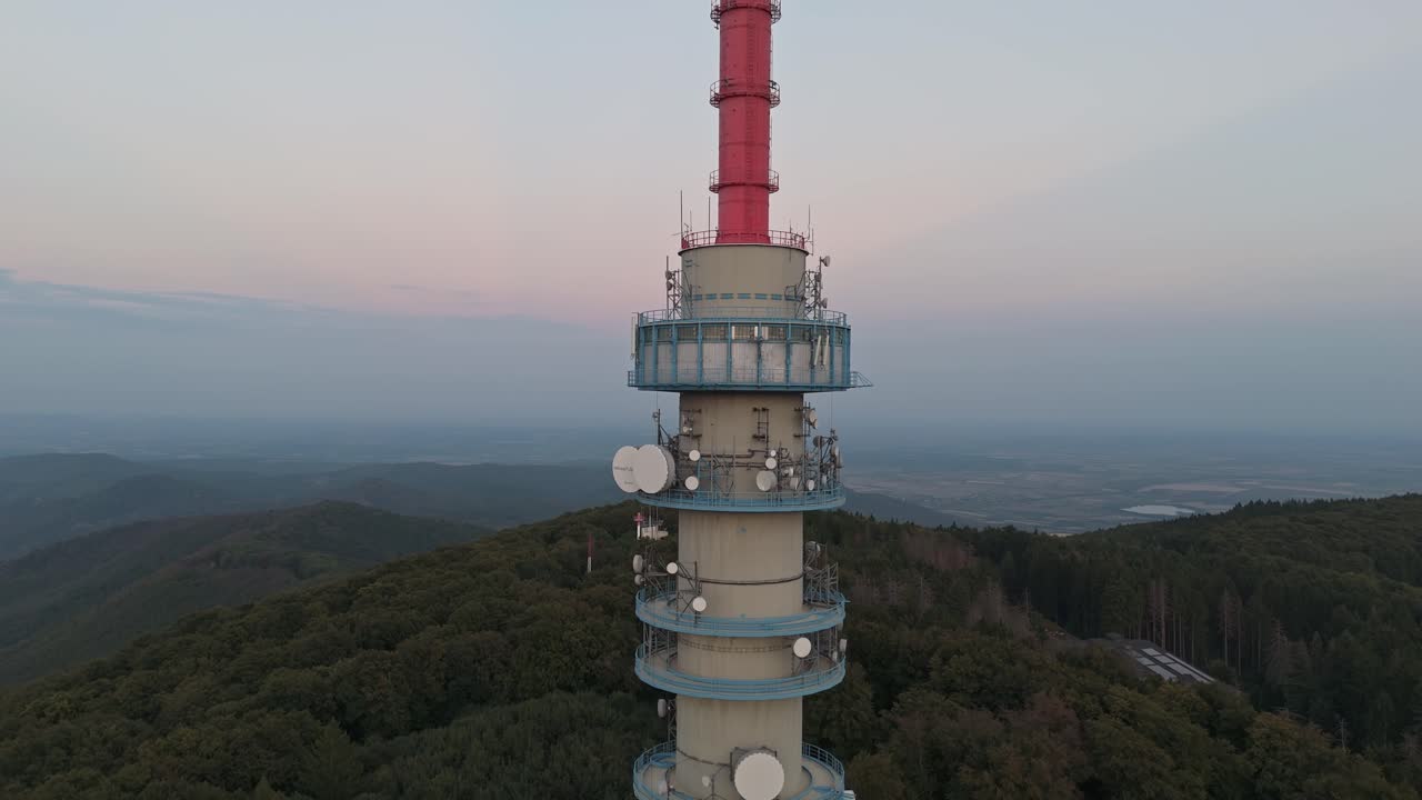 Ascending aerial view along the Kékes TV Tower from the base building towards the top, featuring various antennas with the hazy Mátra Mountains in the background in Hungary