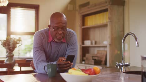 African-american-senior-man-leaning-on-kitchen-counter-using-smartphone-and-smiling