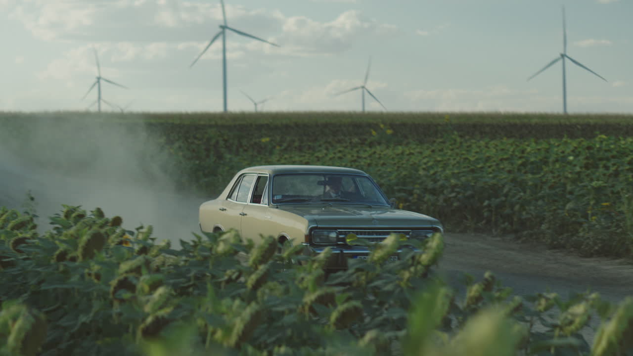 coche de época conduciendo a través de un campo de girasol con turbinas eólicas en el fondo
