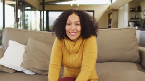 happy mixed race woman sitting on couch in living room, waving and smiling during video call