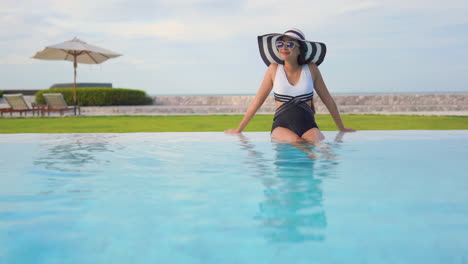 Una-Mujer-Joven,-Con-Un-Traje-De-Baño-De-Una-Pieza-En-Blanco-Y-Negro-Y-Un-Sombrero-Flexible-A-Juego-Para-El-Sol,-Se-Sienta-En-El-Borde-Poco-Profundo-De-La-Piscina-De-Un-Centro-Turístico