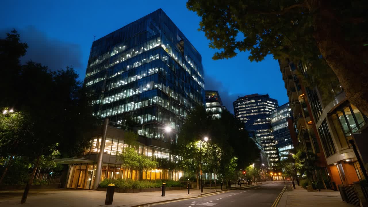 Edifícios urbanos iluminados ao entardecer e à noite com reflexo nas janelas de vidro em uma rua vazia, mostrando a arquitetura moderna e a vida na cidade