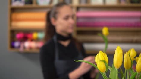 Joven-Atractiva-Mujer-Sonriente-En-Delantal-Trabajando-En-Tiro-Floral-Y-Arreglando-Un-Ramo-De-Flores-Usando-Tulipanes-Amarillos-Frescos