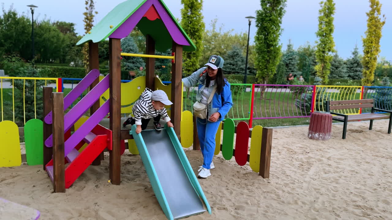 Woman playing with her baby son at playground. Active toddler slides down and crawls by the sand.