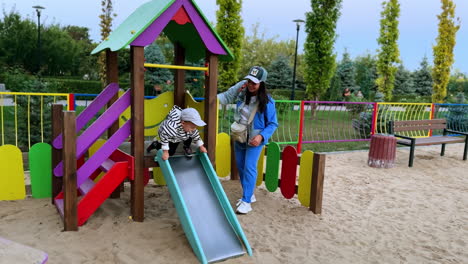 woman playing with her baby son at playground. active toddler slides down and crawls by the sand.