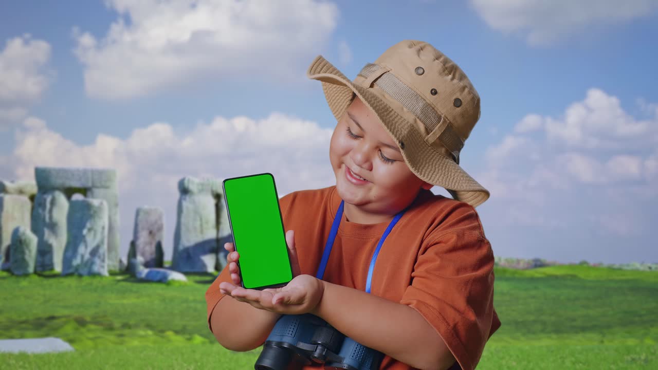 Asian Boy With A Hat And Binoculars Smiling And Showing Green Screen Smartphone To Camera. Boy Researcher Examines Something While Traveling In Stonehenge, Travel Tourism Adventure, Close Up