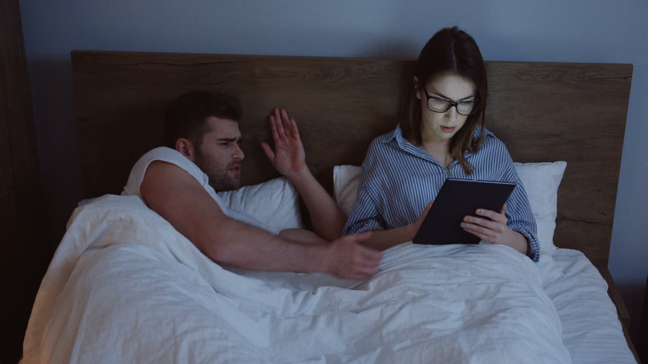 Caucasian young woman in glasses working or chatting on the tablet device late at night while her husband waking up angry and quarreling at her