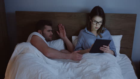mujer joven caucásica con anteojos trabajando o charlando en el dispositivo de la tableta a altas horas de la noche mientras su marido se despierta enojado y peleando con ella