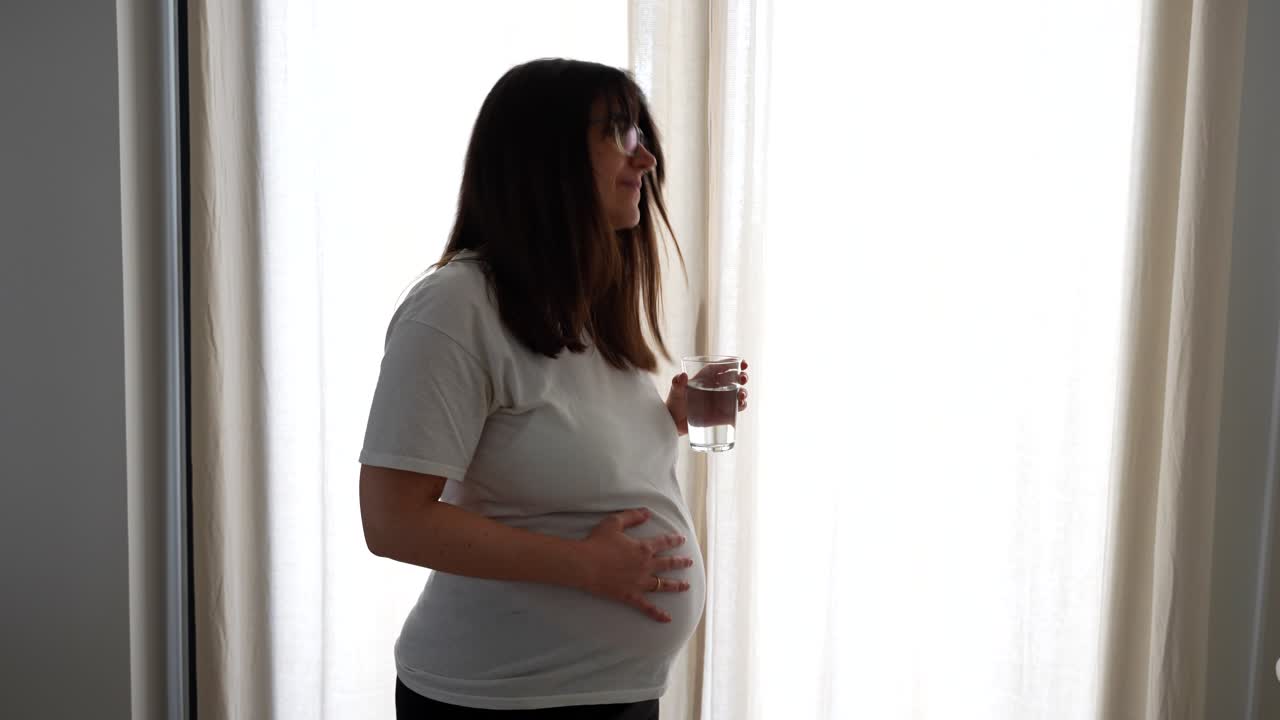 Pregnant woman touches her belly and drinks a glass of water at home, happily looking at the camera