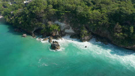 Strong-ocean-waves-crashing-on-tropical-rocky-cliffs.-Aerial-view-of-turquoise-blue-water-splashing-on-caves-and-stone-cliffs-with-rainforest