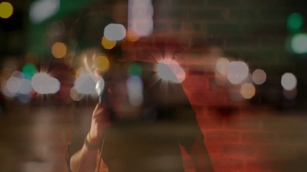 Male performer singing music on city night, holding microphone showing brick overlay, bokeh lights
