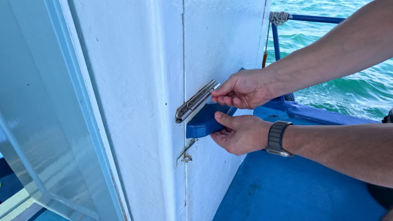 A person unlocks and opens a boat's engine room door on clear waters in Phuket, Thailand. Bright daylight enhances visibility