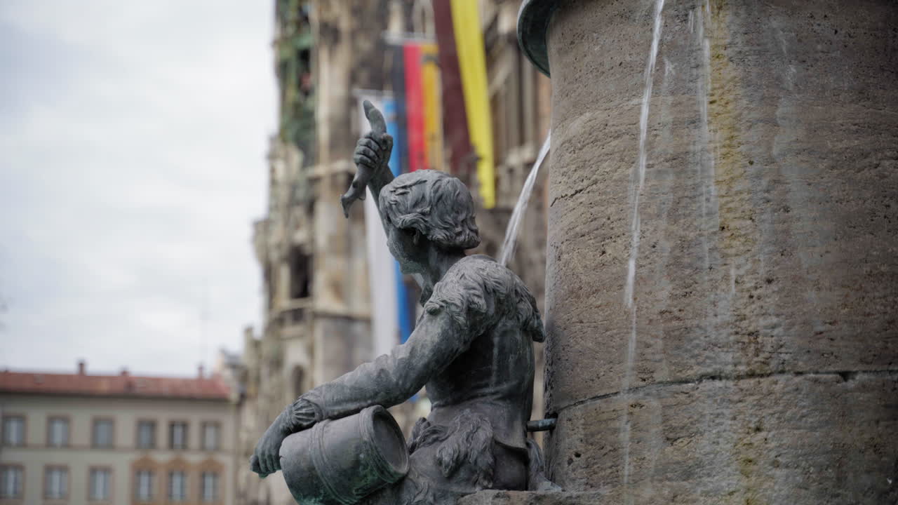 A bronze statue at the Fischbrunnen fountain in Munich’s Marienplatz. The figure holds a fish and a bucket, symbolizing the city's historical traditions. In the background, the Neues Rathaus