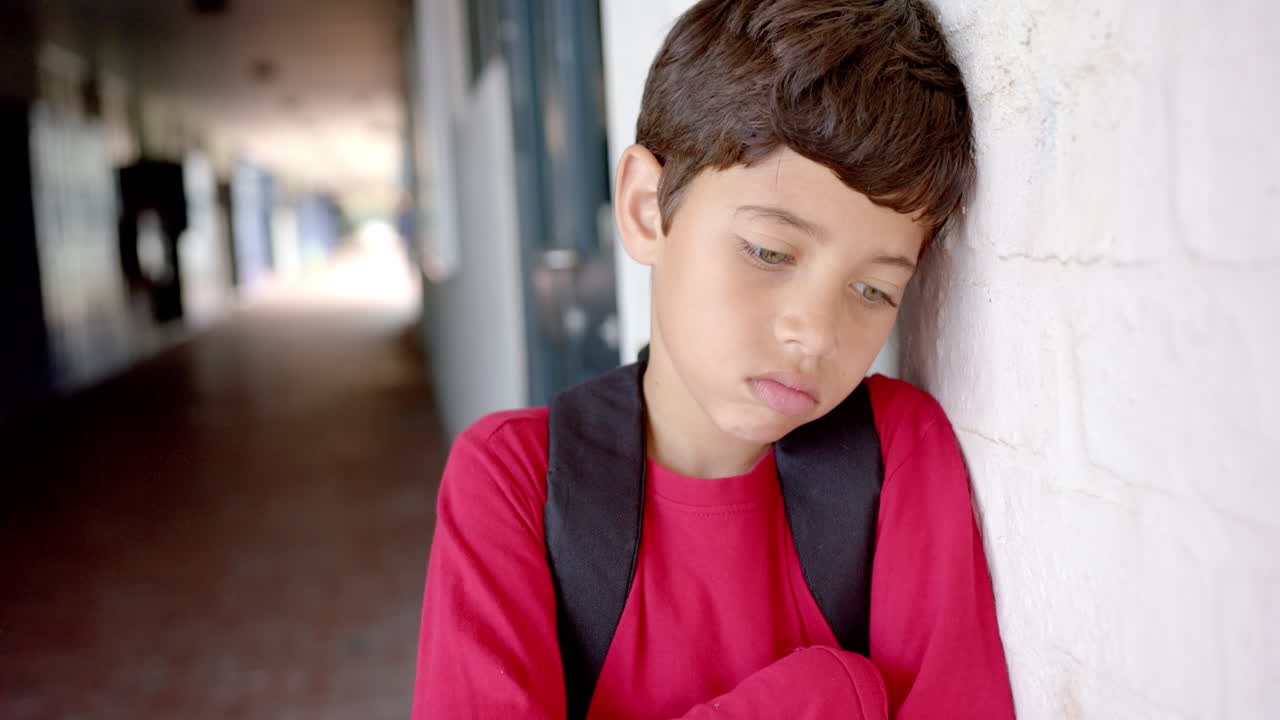 In school, boy with backpack leaning against wall, looking sad and thoughtful