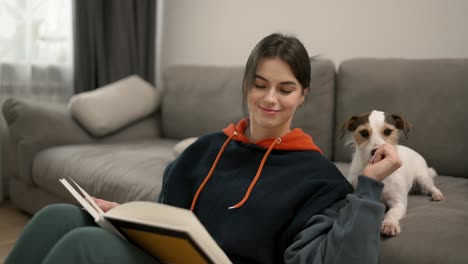 una hermosa mujer leyendo un libro y acariciando a su perro doméstico en el sofá en casa