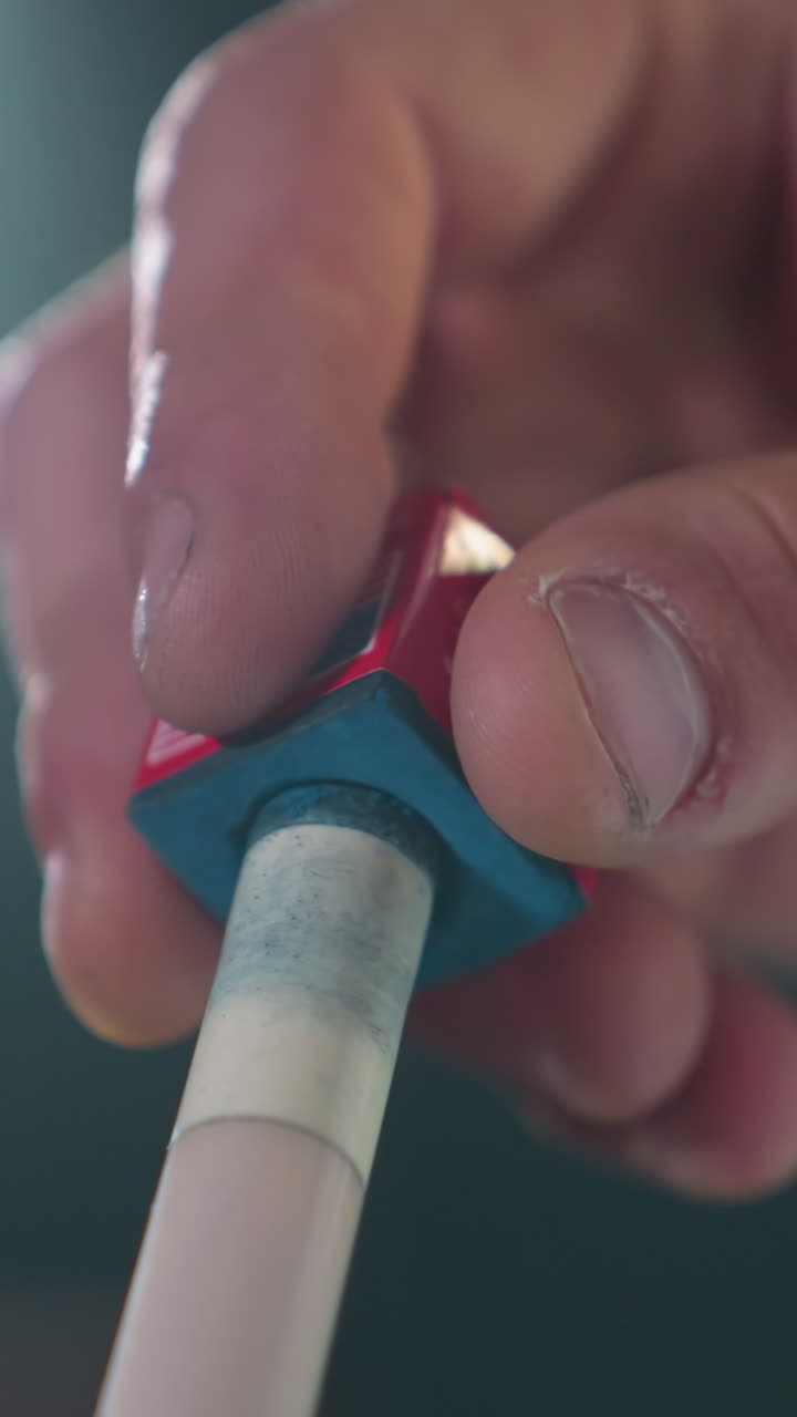 손가락이 조심스럽게 빨간색과 파란색 도자기를 빌리어드 게임 전에 큐 스틱에 적용하는 클로즈업. 집중된 손잡이와 흐릿한 배경은 정확성, 준비 및 주의를 강조합니다.