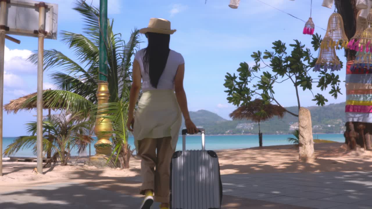 4k donne di viaggio asiatiche che camminano con la sua borsa di viaggio sulla strada laterale alla spiaggia durante una vista di un giorno soleggiato. scattato da dietro. donne turisti felici camminare con le valigie sulla strada della spiaggia. vacanza di viaggio concetto di vacanza estiva.