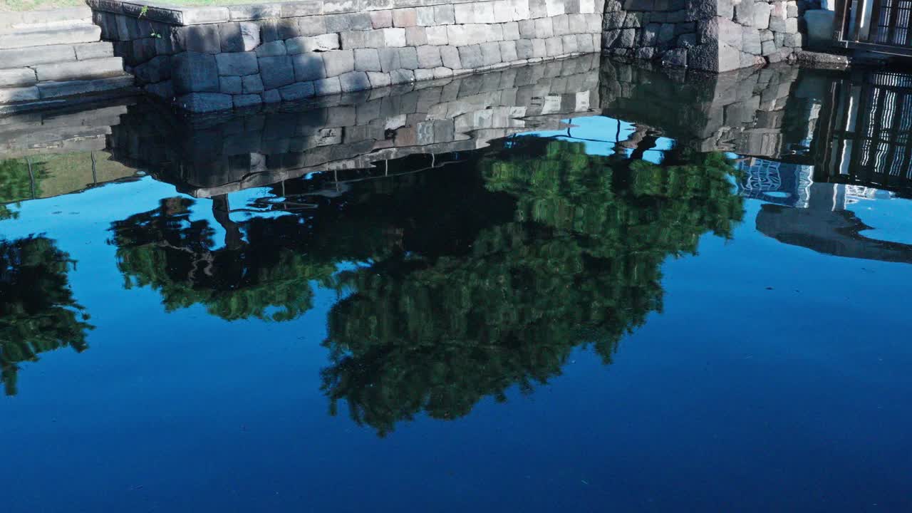 A clear blue pond in Hamarikyu Gardens perfectly reflects a traditional stone bridge and green trees.