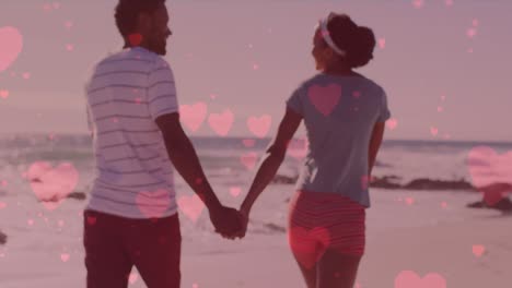multiple pink hearts floating against african american couple holding hands and walking on the beach
