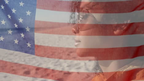 Animation-of-American-flag-waving-over-woman-with-sunglasses-sitting-on-beach-by-seaside