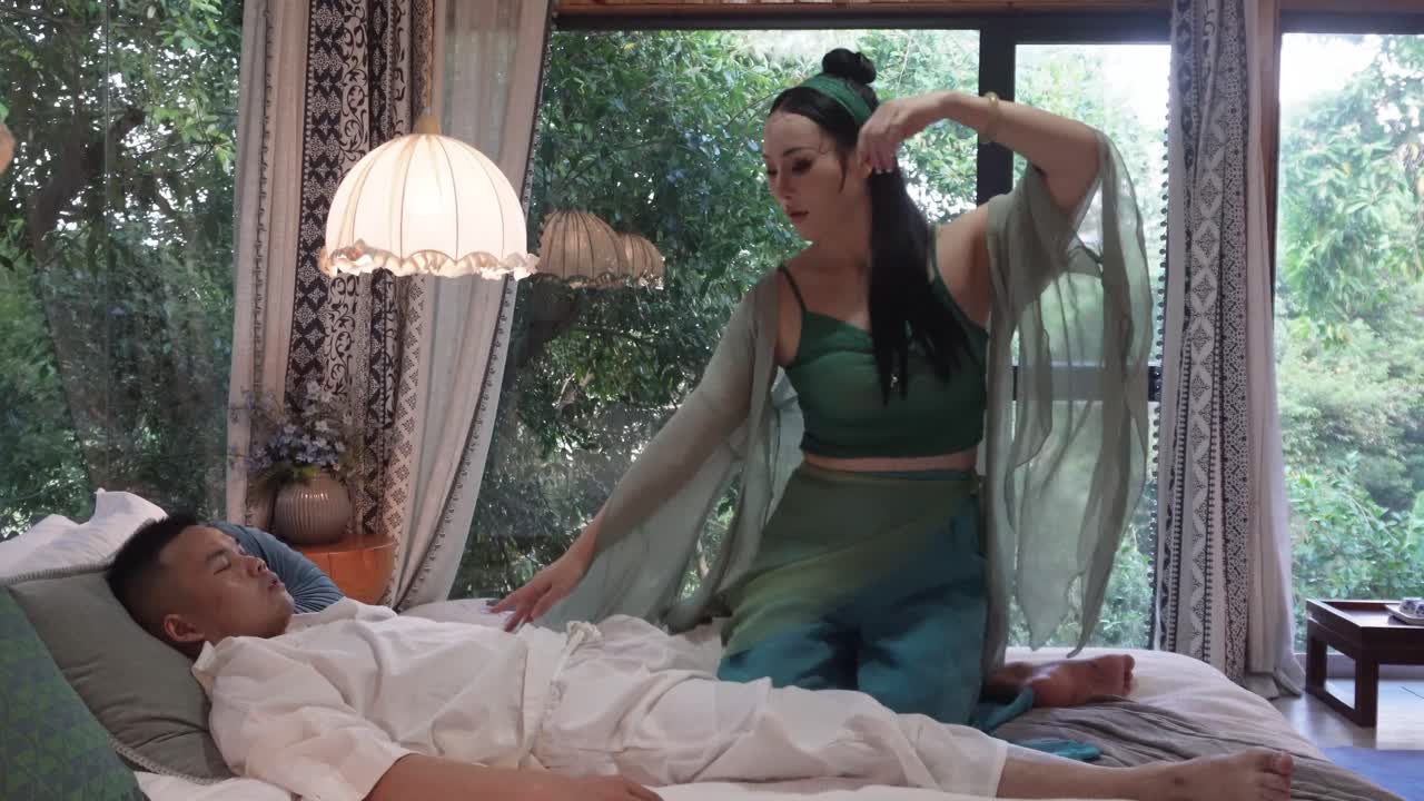 The woman, seated on the bed, performs a ritualistic gesture with her hands, as the man lies motionless beside her